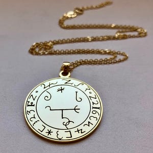 Pode incluir: Colar com pingente dourado com um pingente circular com símbolos e texto pretos. O pingente tem um centro branco e uma borda dourada. A corrente também é dourada. O colar é exibido em uma superfície cinza claro.