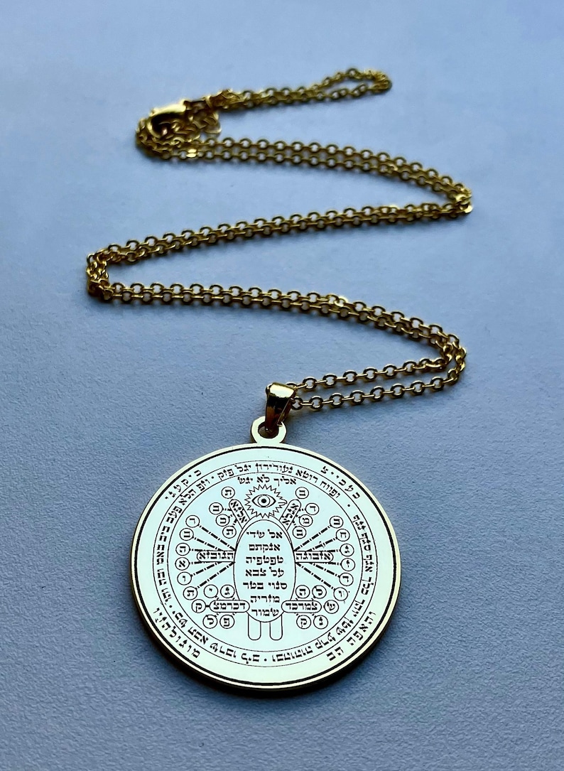 May include: Gold-colored pendant necklace with a circular pendant. The pendant features intricate engraved symbols and text, suspended from a delicate chain. The necklace is displayed against a light gray background.