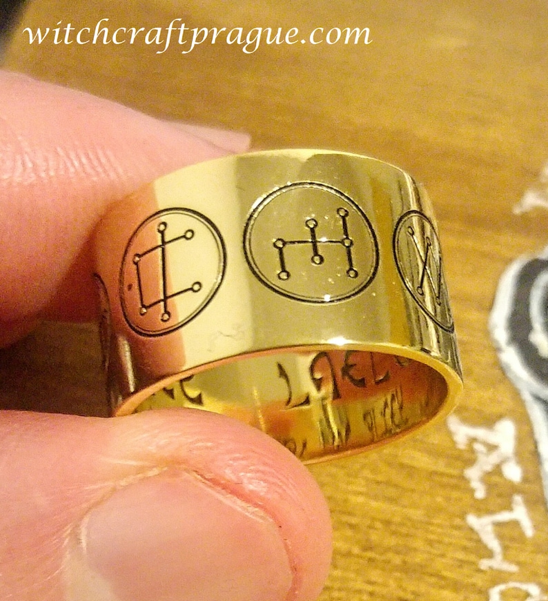 Op de afbeelding: Een gouden ring met vijf gegraveerde symbolen. De symbolen bevinden zich in een cirkel en worden omringd door een gouden band. De ring ligt op een houten oppervlak.