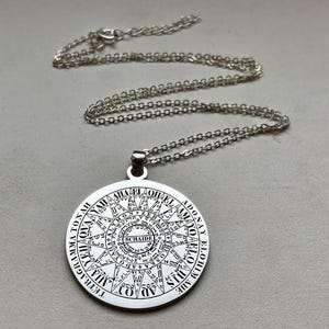 Könnte beinhalten: Eine silberfarbene, runde Anhängerkette mit detaillierten, schwarz gravierten Texten und geometrischen Mustern. Das Design zeigt die Wörter TETRAGRAMMATON, ADONAY, ELOHIM und SCHAIDE, die in konzentrischen Kreisen mit Symbolen angeordnet sind.