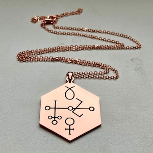 May include: A rose gold-coloured necklace with a hexagonal pendant. The pendant has etched black symbols, possibly astrological or alchemical. The necklace hangs from a fine chain. The necklace is displayed on a light grey surface.