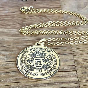 May include: Gold-colored pendant necklace with a circular charm. The charm features intricate symbols and text, with a central design of a hand and eye. The chain is a delicate link style. The background is a wooden surface.