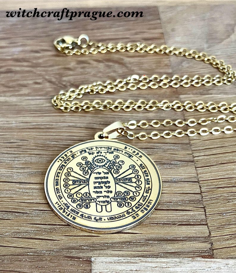 May include: Gold-colored pendant necklace with a circular charm. The charm features intricate symbols and text, with a central design of a hand and eye. The chain is a delicate link style. The background is a wooden surface.