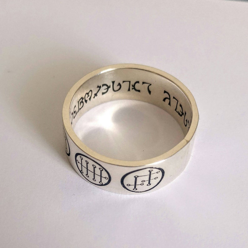 Op de afbeelding: Een zilveren ring met zwarte markeringen. De ring heeft twee ronde ontwerpen met lijnen erin en Hebreeuwse tekst rond de band. De tekst luidt "לְבַדִּי אֱלֹהִים"