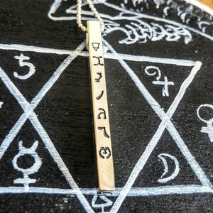 May include: A silver necklace with a long, rectangular pendant. The pendant has a series of symbols engraved on it, including a triangle, a star, and a crescent moon. The necklace is lying on a black surface with a white star of David drawn on it.