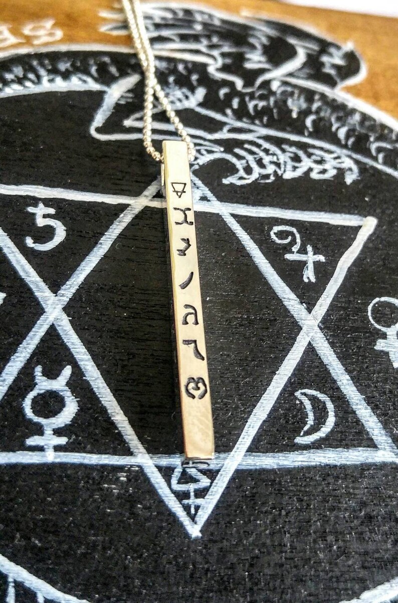 May include: A silver necklace with a long, rectangular pendant. The pendant has a series of symbols engraved on it, including a triangle, a star, and a crescent moon. The necklace is lying on a black surface with a white star of David drawn on it.