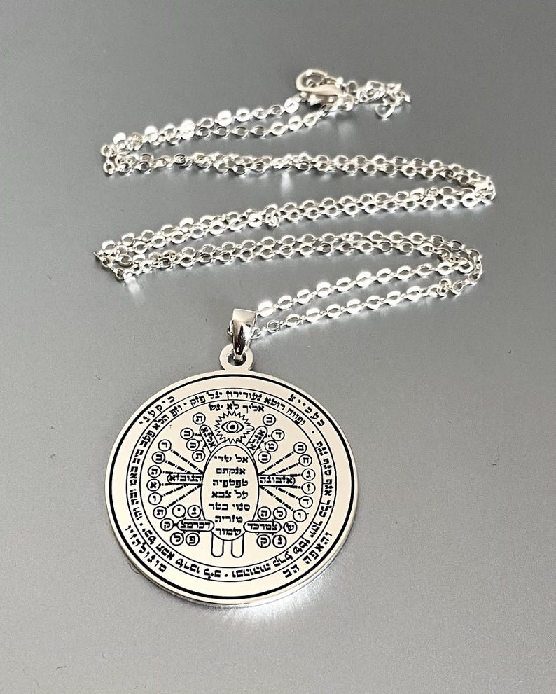 May include: Silver-colored pendant necklace. The pendant is a round disc with intricate engraved symbols and text. The chain is delicate and silver. The pendant has a loop for the chain to pass through.