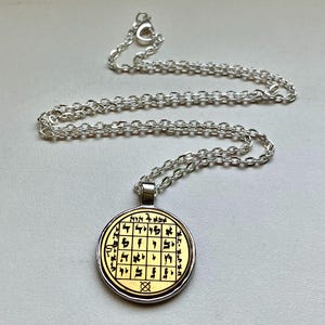 Puede incluir: Collar plateado con un colgante circular. El colgante presenta un centro dorado con símbolos y texto negros, rodeado por un borde plateado. La cadena es de estilo eslabón delicado.