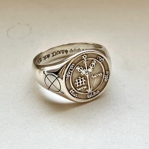 May include: A polished silver signet ring featuring a circular face engraved with intricate occult symbols, Hebrew characters, and a central staff design. The band displays a geometric cross-circle emblem on the side and additional symbols inside.