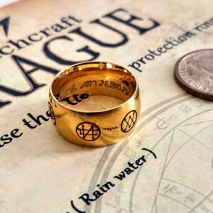 Op de afbeelding: Een gouden ring met een zwarte inscriptie en symbolen. De ring ligt op een boek met de zichtbare woorden "Witchcraft Prague" en "protection ring".