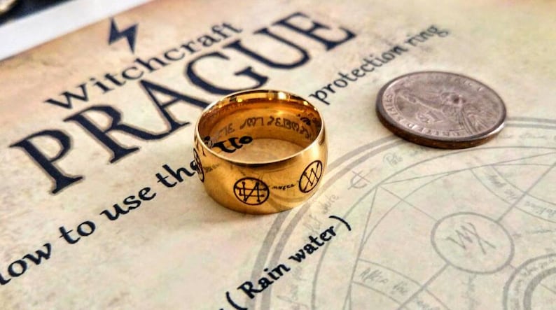 Op de afbeelding: Een gouden ring met een zwarte inscriptie en symbolen. De ring ligt op een boek met de zichtbare woorden "Witchcraft Prague" en "protection ring".