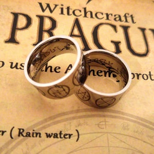 Op de afbeelding: Twee zilveren ringen met zwarte inscripties en symbolen. De ringen liggen op een bruine achtergrond met de tekst "Witchcraft Prague" en "Hoe te gebruiken de... bescherming"