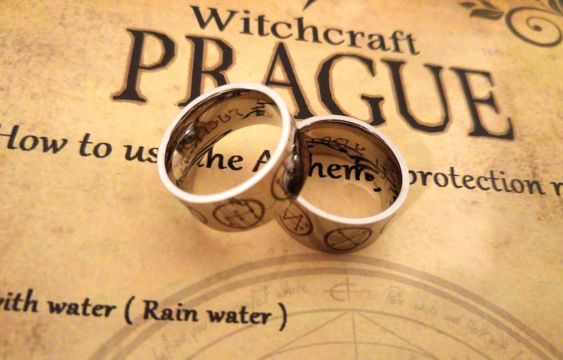 Op de afbeelding: Twee zilveren ringen met zwarte inscripties en symbolen. De ringen liggen op een bruine achtergrond met de tekst "Witchcraft Prague" en "Hoe te gebruiken de... bescherming"