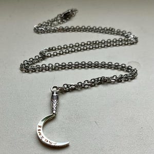 Op de afbeelding: Zilverkleurige ketting met een halve maanvormige hanger met geëtste letters. De hanger is bevestigd aan een fijne ketting. De ketting heeft een gedetailleerd ontwerp en is waarschijnlijk gemaakt van metaal.