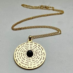 Puede incluir: Un collar con colgante circular en tono dorado que presenta una piedra negra central rodeada por anillos concéntricos de texto hebreo grabado. El colgante cuelga de una delicada cadena de eslabones redondos dorada con cierre de mosquetón.