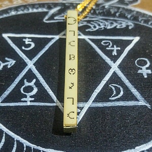 May include: A gold bar necklace with a series of symbols engraved on it, including a crescent moon, a star, a heart, and a cross. The necklace is hanging over a black surface with a white pentagon drawn on it.