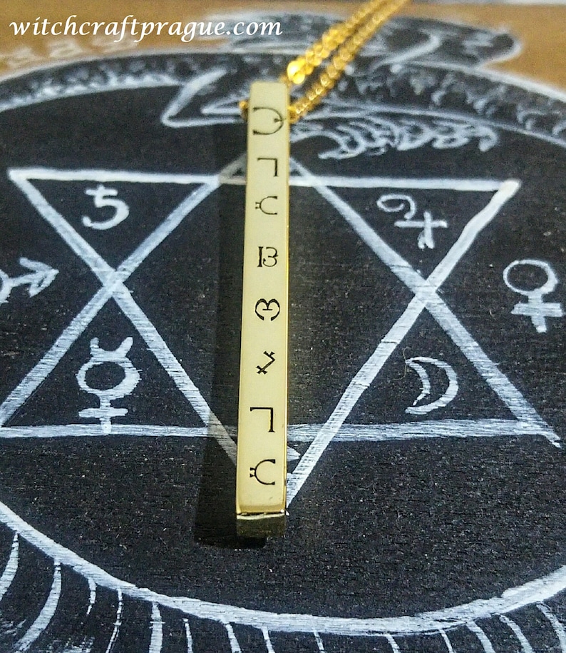 May include: A gold bar necklace with a series of symbols engraved on it, including a crescent moon, a star, a heart, and a cross. The necklace is hanging over a black surface with a white pentagon drawn on it.