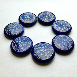 Puede incluir: Siete piedras circulares de color azul intenso pulidas y dispuestas en círculo. Cada piedra presenta intrincados grabados dorados de símbolos místicos, líneas geométricas y letras como A, N y Z, junto con palabras como AGLA y SADAY.