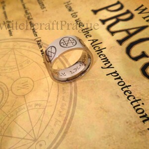 Op de afbeelding: Een zilveren ring met een inscriptie en een witte achtergrond met verschillende symbolen. De ring ligt op een stuk papier met de tekst "Witchcraft Prague How to use the Alchemy protection ring".