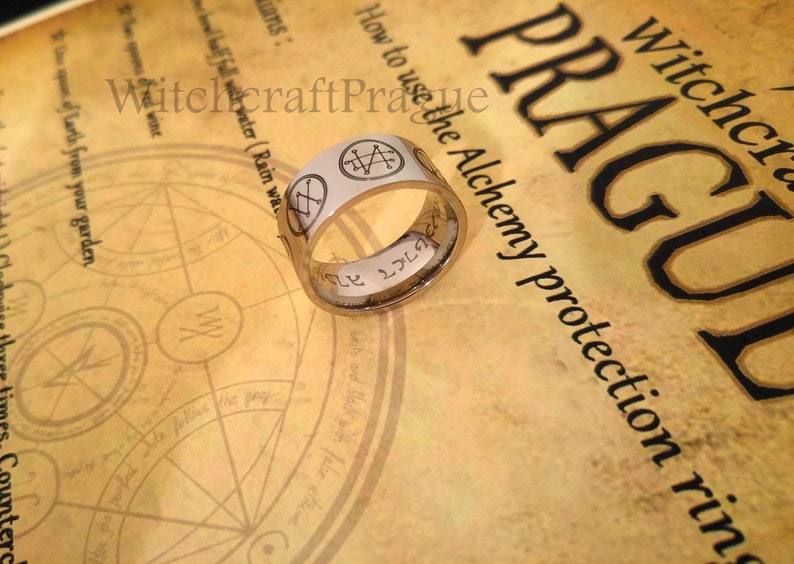 Op de afbeelding: Een zilveren ring met een inscriptie en een witte achtergrond met verschillende symbolen. De ring ligt op een stuk papier met de tekst "Witchcraft Prague How to use the Alchemy protection ring".