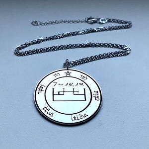 Könnte beinhalten: Eine silberfarbene Anhängerkette mit einem kreisförmigen Anhänger. Der Anhänger hat eingravierte Symbole und Text am Rand und in der Mitte. Die Halskette hat eine filigrane Gliederkette.