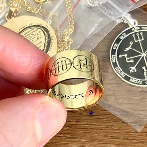 May include: A wide gold-toned band ring featuring engraved black circular sigil symbols on the exterior surface. The interior of the ring is also engraved with black lettering. The ring is positioned in front of two circular pendants in plastic bags.