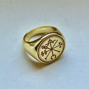 May include: A polished gold signet ring featuring a circular face with an engraved occult-style sigil. The design consists of intersecting lines and various geometric symbols, including a crescent, a cross, and loops, centered on the band.
