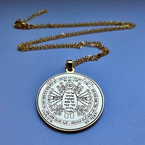 May include: A gold-colored pendant necklace with a circular charm. The charm features intricate text and symbols in a circular pattern. The chain and pendant are a shiny gold color, set against a blue background. The necklace is displayed from above.