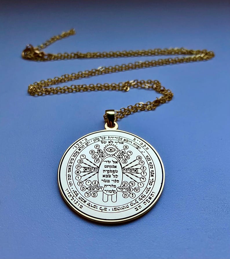 May include: A gold-colored pendant necklace with a circular charm. The charm features intricate text and symbols in a circular pattern. The chain and pendant are a shiny gold color, set against a blue background. The necklace is displayed from above.