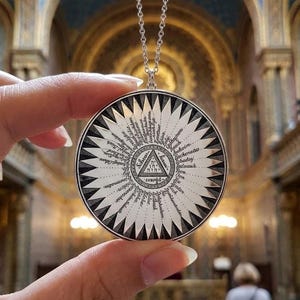 May include: A circular silver pendant on a delicate chain featuring a central triangle with Hebrew text and a surrounding sunburst pattern. The design includes radiating lines and various words inscribed in black ink against a white background.