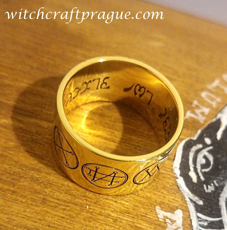 Op de afbeelding: Een gouden ring met zwarte gegraveerde symbolen en de tekst "L97 3LXC" aan de binnenkant van de band.