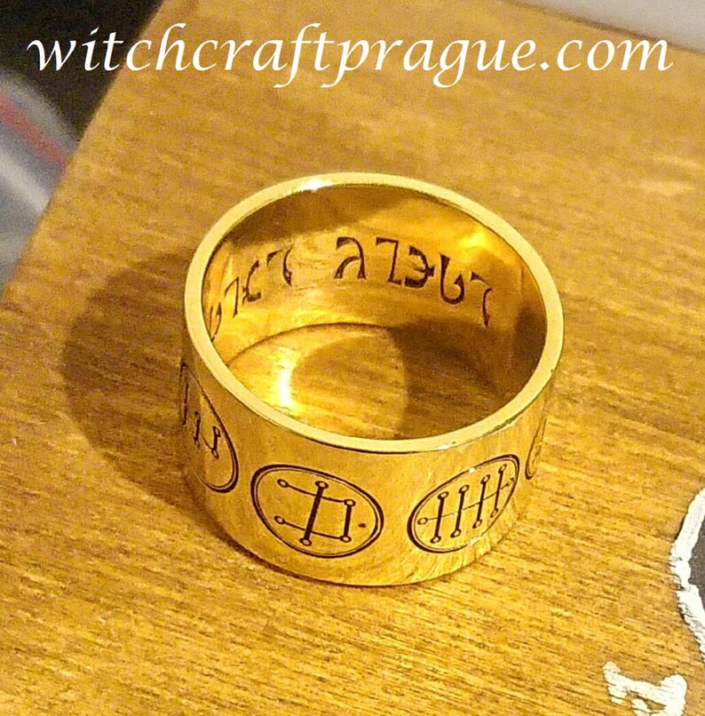 Op de afbeelding: Een gouden ring met gegraveerde symbolen en tekst. De tekst op de ring luidt "witchcraftprague.com".