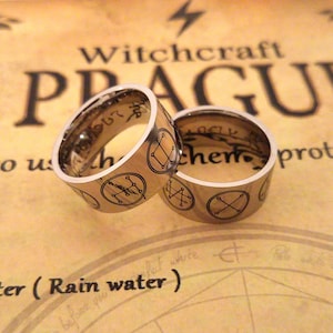 Op de afbeelding: Twee zilveren ringen met gegraveerde symbolen en de woorden "Witchcraft Prague" en "alchemie" op een bruine achtergrond.