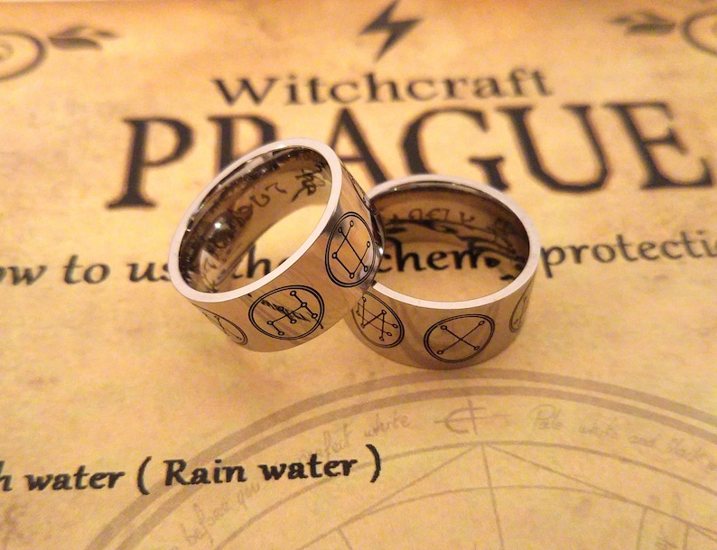 Op de afbeelding: Twee zilveren ringen met gegraveerde symbolen en de woorden "Witchcraft Prague" en "alchemie" op een bruine achtergrond.