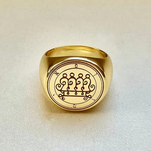 Könnte beinhalten: Ein goldfarbener Siegelring mit einem kreisförmigen Emblem. Das Emblem zeigt ein kompliziertes Design mit stilisierten Figuren und Symbolen. Der Ring hat eine polierte Oberfläche und ein abgerundetes Band.