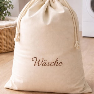 Könnte beinhalten: Ein beiger Wäschebeutel mit Kordelzug. Auf dem Beutel ist das Wort "Wäsche" in brauner Schrift gestickt. Der Beutel besteht aus einem weichen, strukturierten Stoff und steht auf einem Holzboden.
