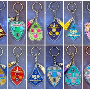 May include: Twelve clear acrylic keychains with a colorful Ouija board design. Each keychain features a different zodiac sign, including Aries, Taurus, Gemini, Cancer, Leo, Virgo, Libra, Scorpio, Sagittarius, Capricorn, Aquarius, and Pisces. Each keychain has a gold-colored key ring and a tassel.
