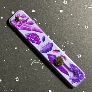May include: Purple and white phone grip with a crystal design. The grip has a gold button.