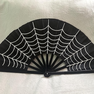 May include: A black folding hand fan featuring a white spider web pattern printed across the fabric. The fan has black ribs and a central pivot point, displaying a semi-circular shape when fully opened to show the complete web design.