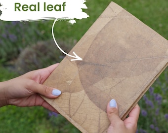 May include: A rectangular notebook with a tan cover featuring a large, pressed real leaf pattern. The cover displays visible organic veins and textures from the leaf. A white graphic overlay in the upper left corner contains the text Real leaf.