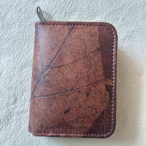 Puede incluir: Una cartera de cuero marrón con diseño de hoja. La cartera rectangular tiene esquinas redondeadas y cierre de cremallera. El patrón de hoja es visible en la parte delantera.
