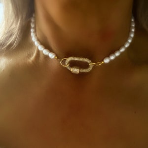 May include: A white pearl necklace with a gold clasp featuring a row of small clear stones. The necklace is worn by a person.