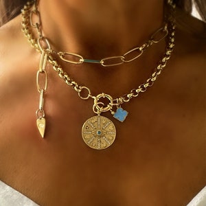 May include: A gold chain necklace with a large round pendant featuring a detailed design with a blue centre. The pendant is attached to a smaller charm shaped like a four-leaf clover. The necklace is layered with a second gold chain with a heart-shaped charm.