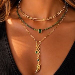 May include: A layered necklace set featuring a gold chain, a strand of small green beads with a central green cylindrical bead, and a gold chain with a dangling gold horn pendant accented by two small green circular stones and a gold link.