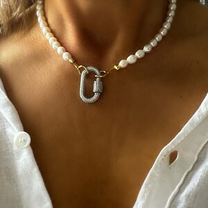 May include: A white pearl necklace with a gold and silver clasp. The clasp is a large, oval-shaped carabiner with clear rhinestones.
