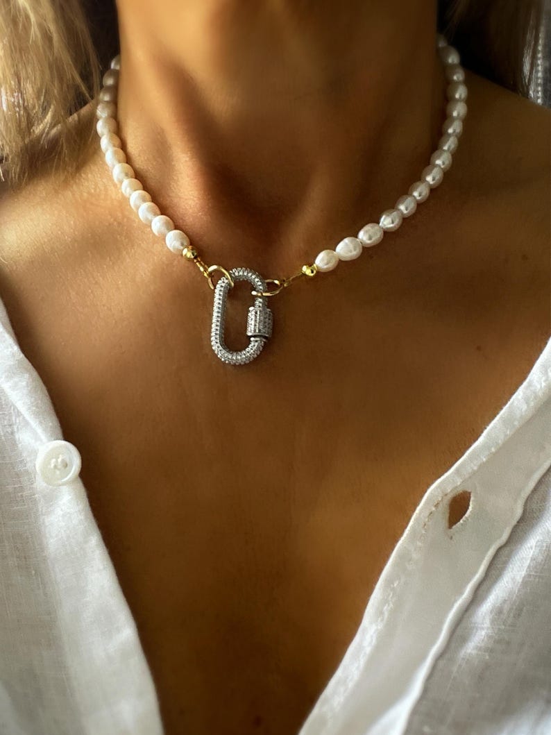 May include: A white pearl necklace with a gold and silver clasp. The clasp is a large, oval-shaped carabiner with clear rhinestones.