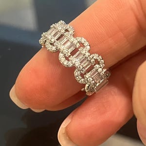 May include: A silver ring with a repeating pattern of baguette-cut and round-cut diamonds. The ring is held between two fingers, showcasing the intricate design and sparkle of the gemstones. The ring is a piece of jewelry.