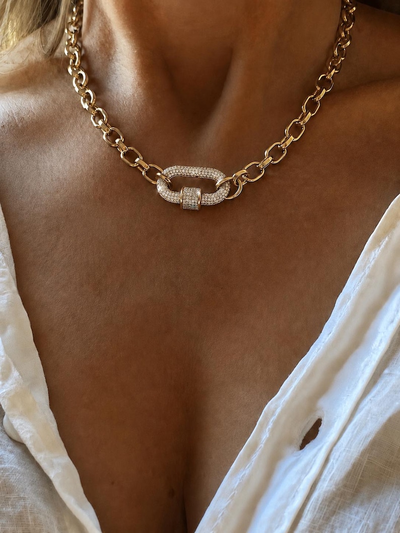Pu&ograve; includere: Collana a catena color oro con un grande pendente ovale. Il pendente e la chiusura sono incastonati con piccoli cristalli scintillanti. La collana &egrave; indossata su una camicia bianca.