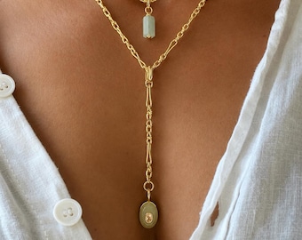 May include: A gold-toned layered chain necklace featuring two distinct pendants. The upper chain holds a small, cylindrical, pale green stone, while the lower chain extends into a Y-shape, ending in an oval, pale green stone with a central gem.