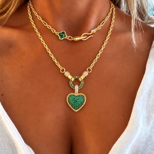 Könnte beinhalten: Eine goldfarbene, mehrlagige Halskette mit einem herzförmigen Anhänger, der mit kleinen grünen Steinen und klaren Kristallen besetzt ist. Die Kette enthält ein kleeblattförmiges grünes Steinelement und einen Hakenverschluss mit einem kleinen Kristall.